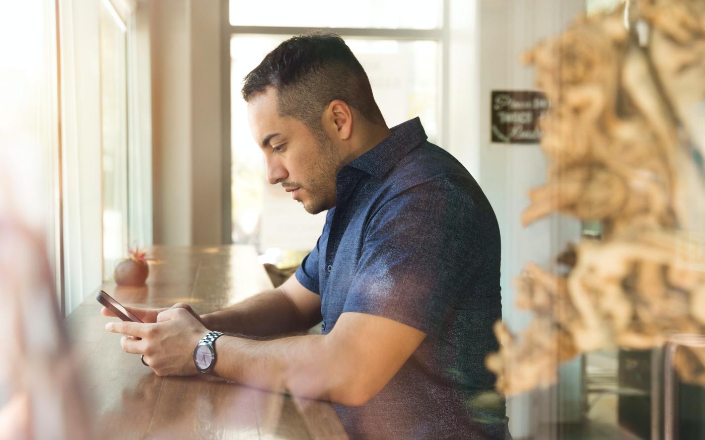 Man sitting at a long table in front of a window. He is looking at his phone.